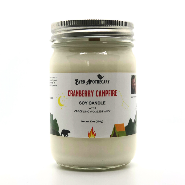 Photo of Byrd Apothecary Cranberry Campfire Soy Candle with crackling Wooden Wick in a canning jar. The label background is a cartoon forest scene with mountains, a walking black bear, a tent with a campfire, a forest and sky with moon, stars and the Big Dipper.